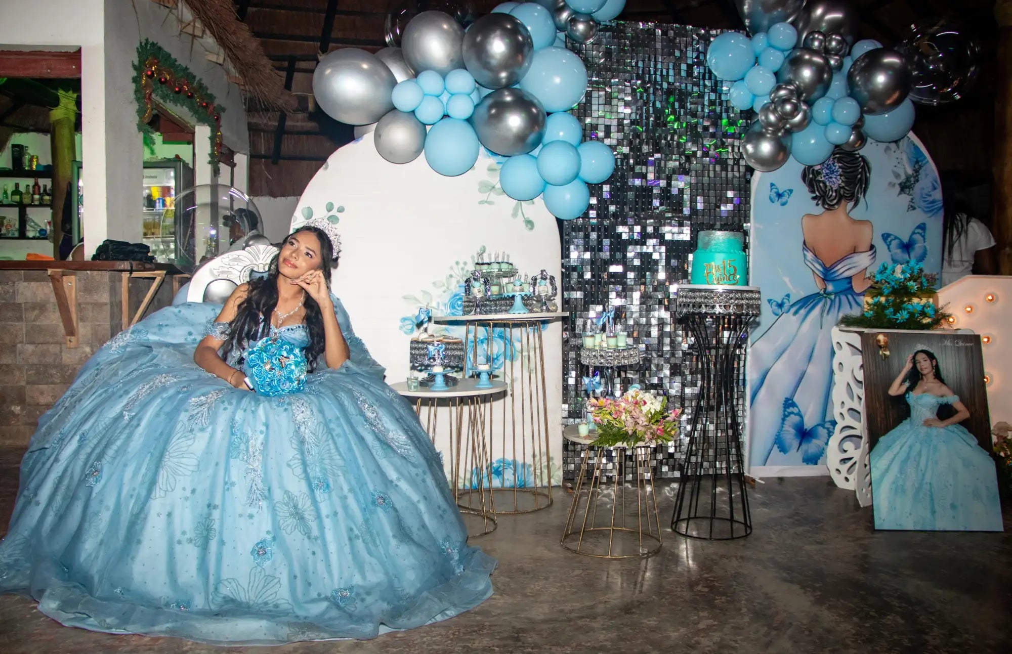 Una mujer en un vestido de quinceañera azul claro, voluminoso, con detalles intrincados de encaje y bordado floral, se sienta con gracia en el centro de una escena de fiesta elaboradamente decorada.